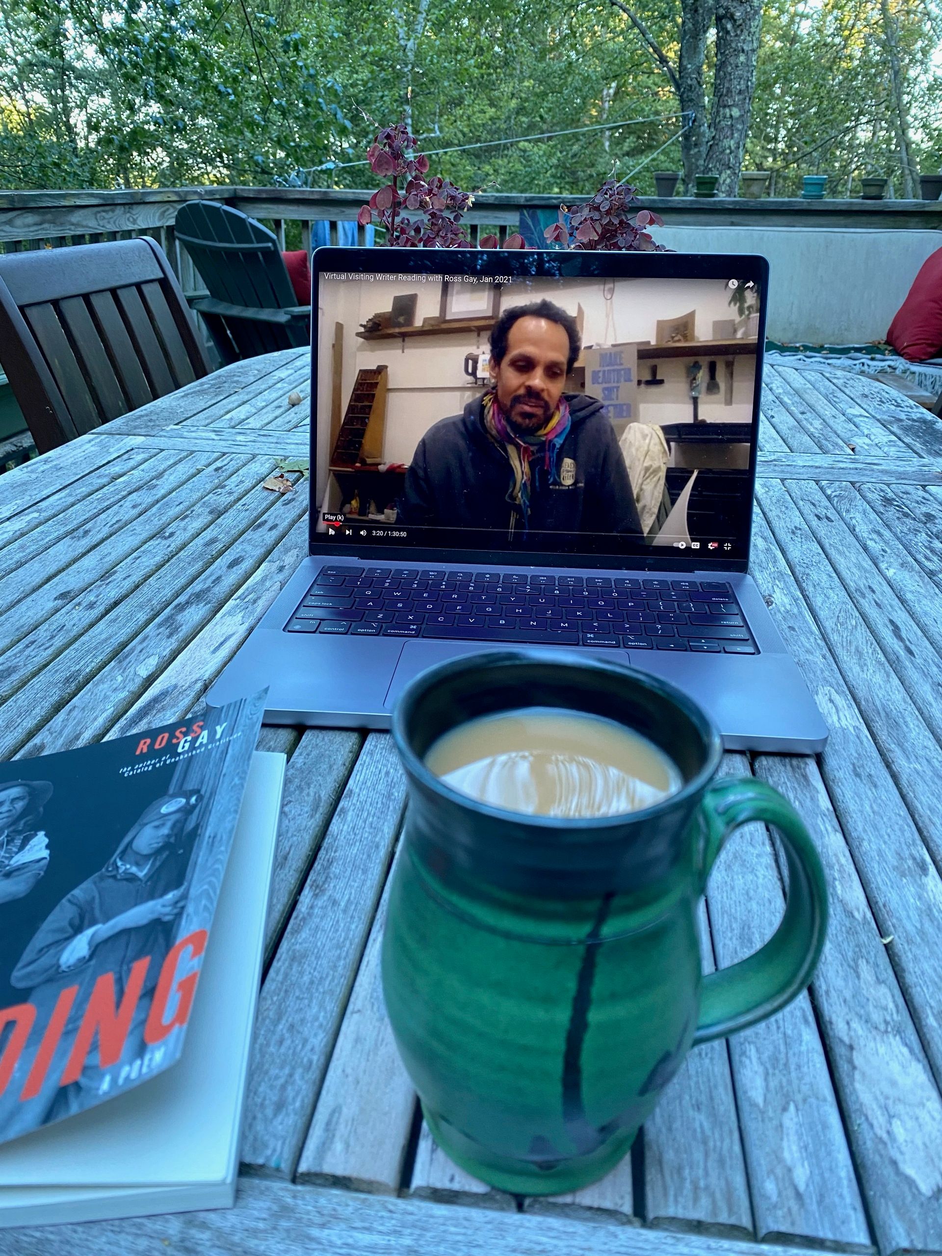 My computer, showing Ross Gay’s face, on a porch table next to a mug of tea and the book Be Holding.