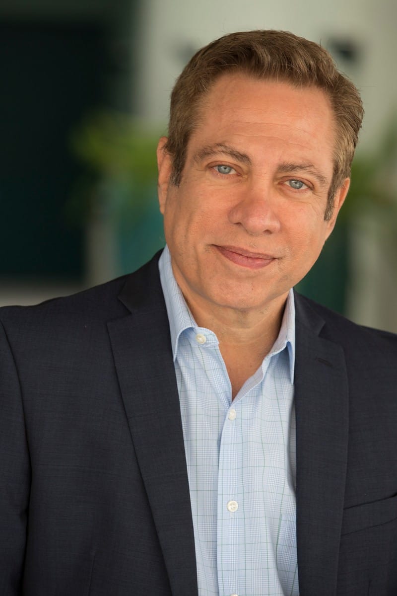 "Portrait of grief expert David Kessler, wearing a dark blazer over a light blue checkered shirt, looking gently into the camera with a slight, compassionate smile. The background is softly blurred, with hints of greenery."