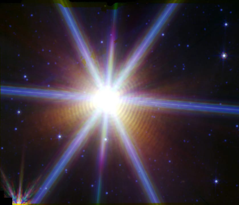 A brilliant star in the center has six bright spikes of light going through it. It’s surrounded by over a dozen thin, symmetric shells of dust colored orange.
