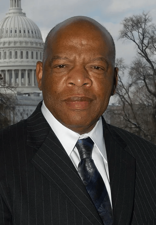 John Lewis, a bald Black man wearing a dark pinstripe suit, white shirt, and patterned blue tie, stands in front of the U.S. Capitol building, looking directly at the camera with a solemn expression. Leafless trees frame the background under a cloudy sky.