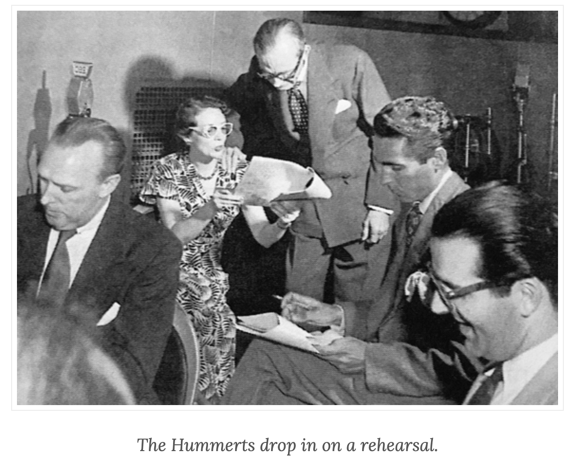 The Hummerts Drop In on a Rehearsal: photo shows a woman in a print dress ad white-framed glasses holding a script and talking with a tall man bending over her in a busy radio studio.