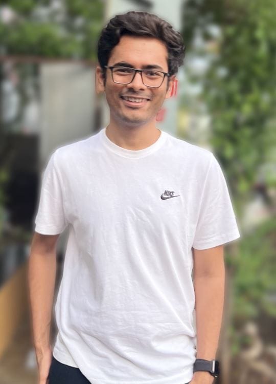 A young man with glasses and short dark hair smiles at the camera. He wears a white Nike t-shirt and a black wristwatch. The background features a blurred outdoor setting with green foliage and a building, suggesting a casual, natural environment.
