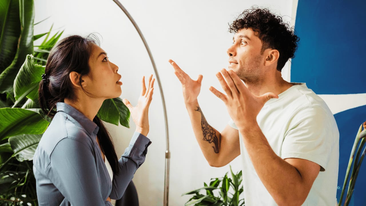Two people standing indoors in a heated argument, both gesturing with raised hands and intense expressions, surrounded by green plants.