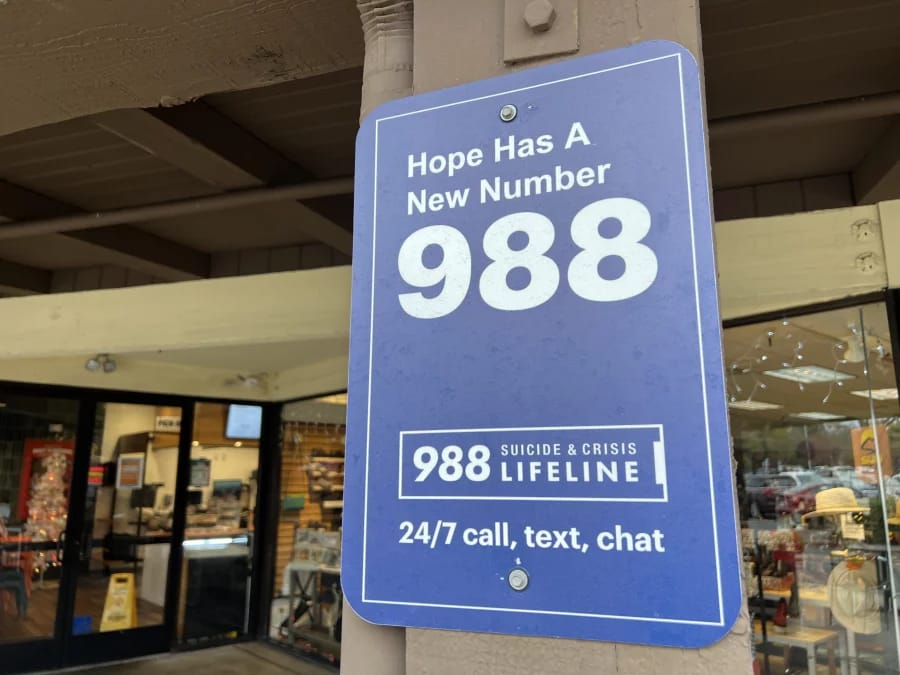 A blue sign posted on a wooden pillar outside a building reads, āHope Has A New Number 988 ā Suicide & Crisis Lifeline ā 24/7 call, text, chat.ā The sign promotes the 988 mental health crisis hotline.