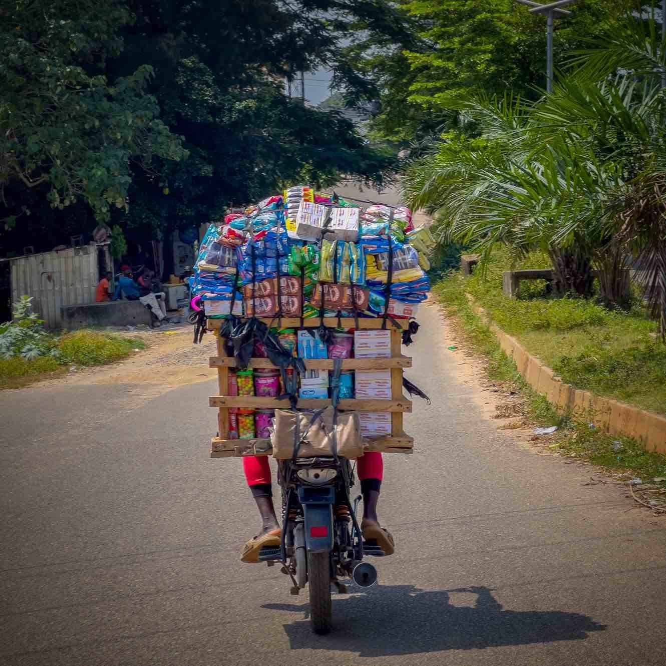 A motorbike rider navigates the edge of Abuja, Nigeria’s capital, carrying a towering load of fast-moving consumer goods including milk, toothpaste, and toothbrushes, supplying makeshift neighborhood stores and showcasing the industrious spirit of everyday Nigerians.