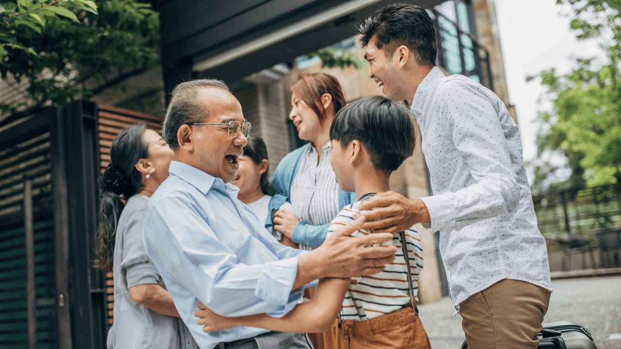 Joyful multigenerational family greeting each other outside, with an older man laughing and hugging a young boy while others smile nearby.