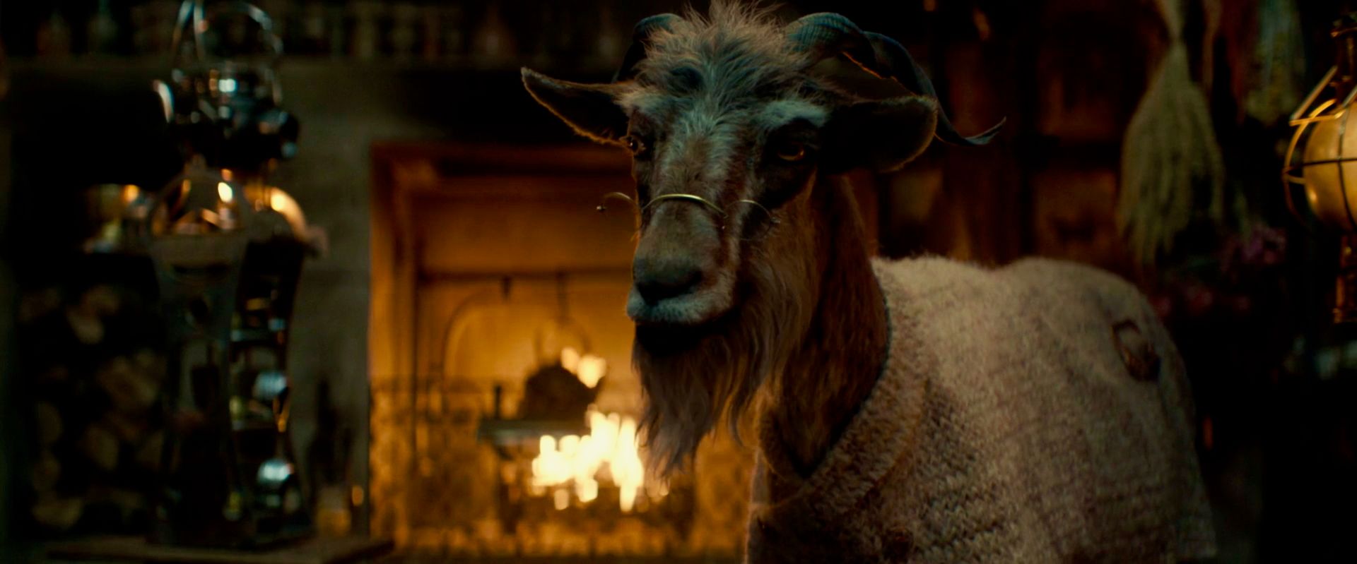 Dr. Dillamond, a goat, is wearing spectacles and an off-white sweater draped over his body in WICKED. He's standing in front of his fireplace.