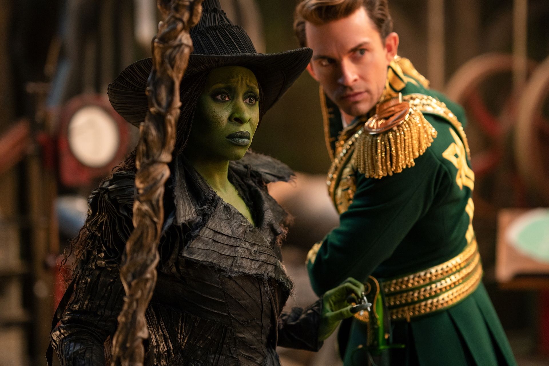 Cynthia Erivo as Elphaba (left) and Jonathan Bailey as Fiyero (right) in WICKED: FOR GOOD. Elphaba is wearing a black outfit, black pointed hat, and is holding a broom while Fiyero is wearing a green military uniform with gold regalia across it.