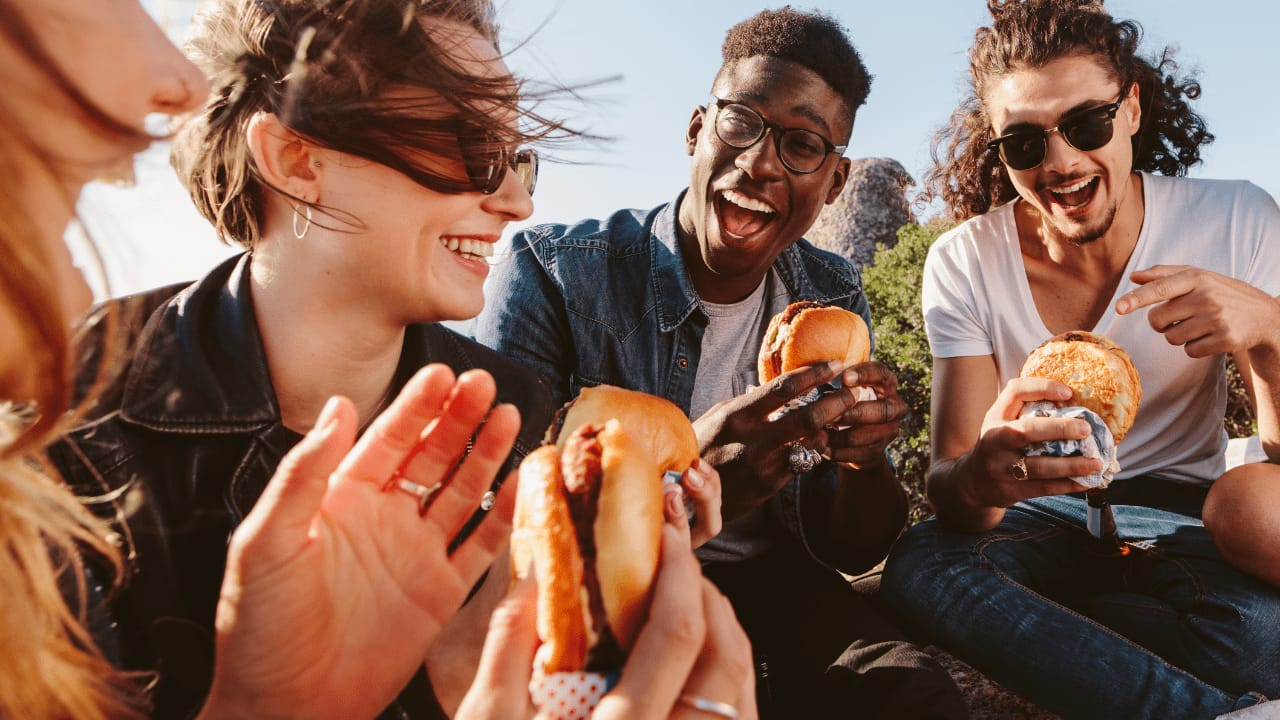 Group of friends laughing and enjoying burgers together outdoors on a sunny day.