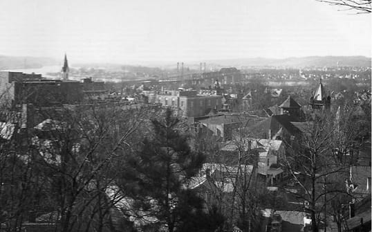 View overlooking a small town, with trees in the foreground, and a bridge, church steeple, and houses in the distance.