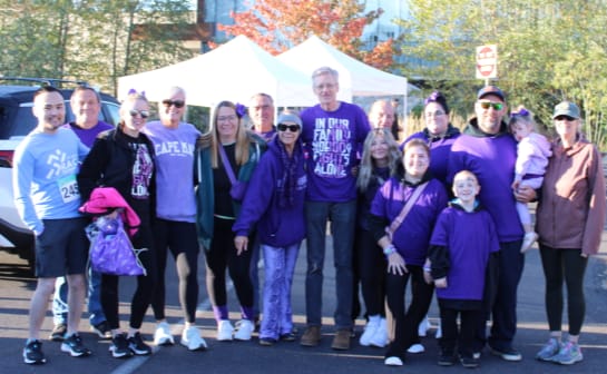 A group of runners and supporters dressed in purple, smiling and posing together at a race event with tents and fall trees in the background.