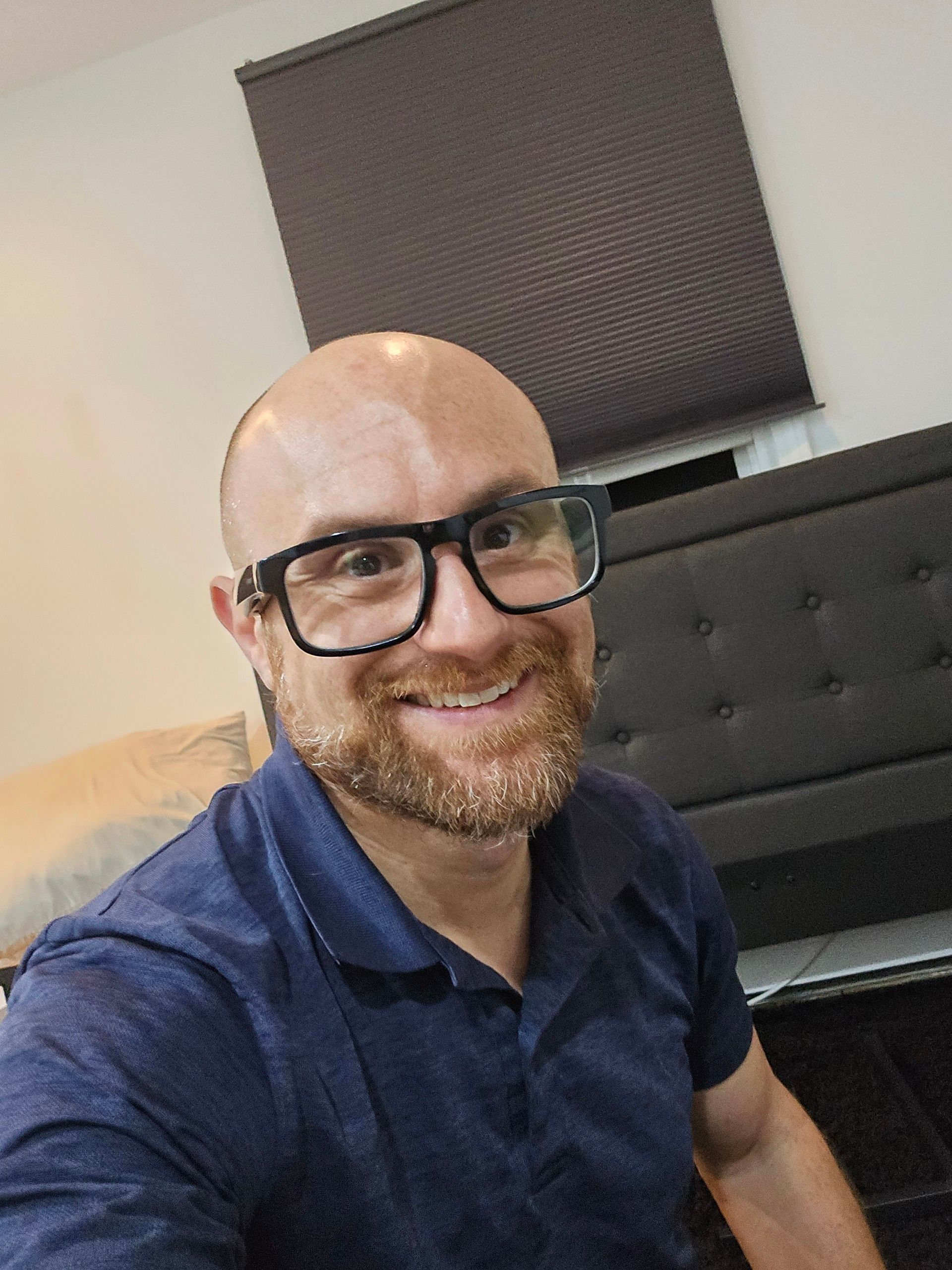 A selfie of me in the middle of a partially assembled bed. I'm a bald, middle-aged, white man with a red beard flecked with white. I'm wearing glasses with thick black rims and a blue collared shirt.
