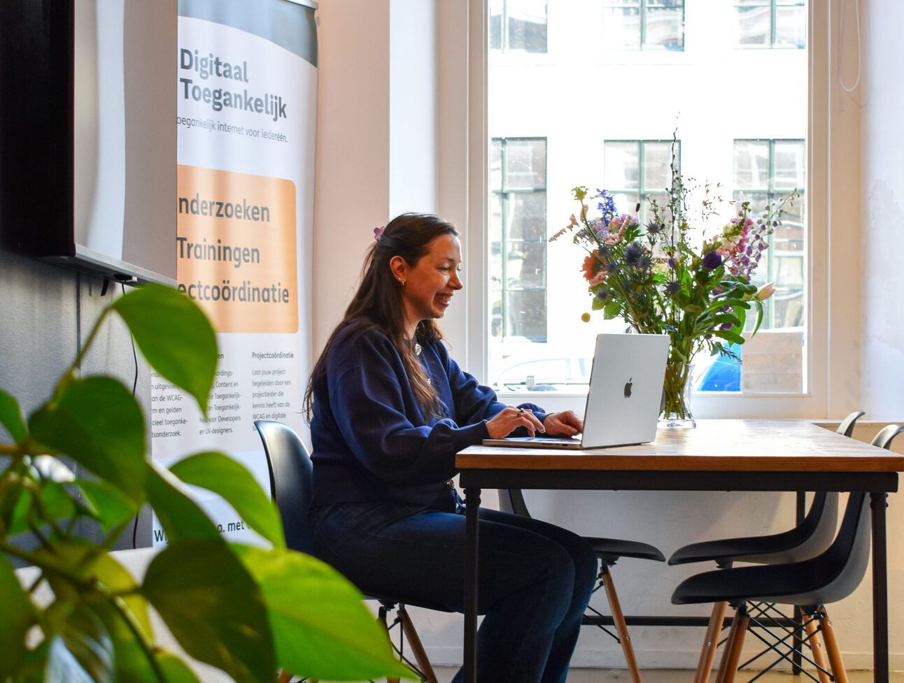 Talissa zit lachend achter haar laptop op het oude kantoor van Digitaal Togankelijk. Achter haar zie je en poster van Digitaal Toegankelijk.
