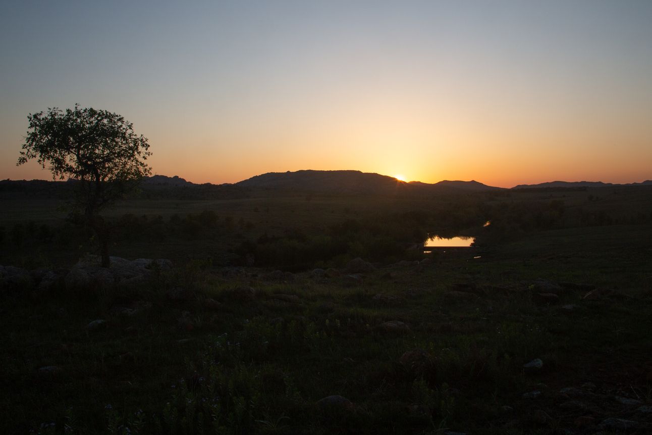 The sun setting over hills in the background, reflected in a body of water in the middle of the image.