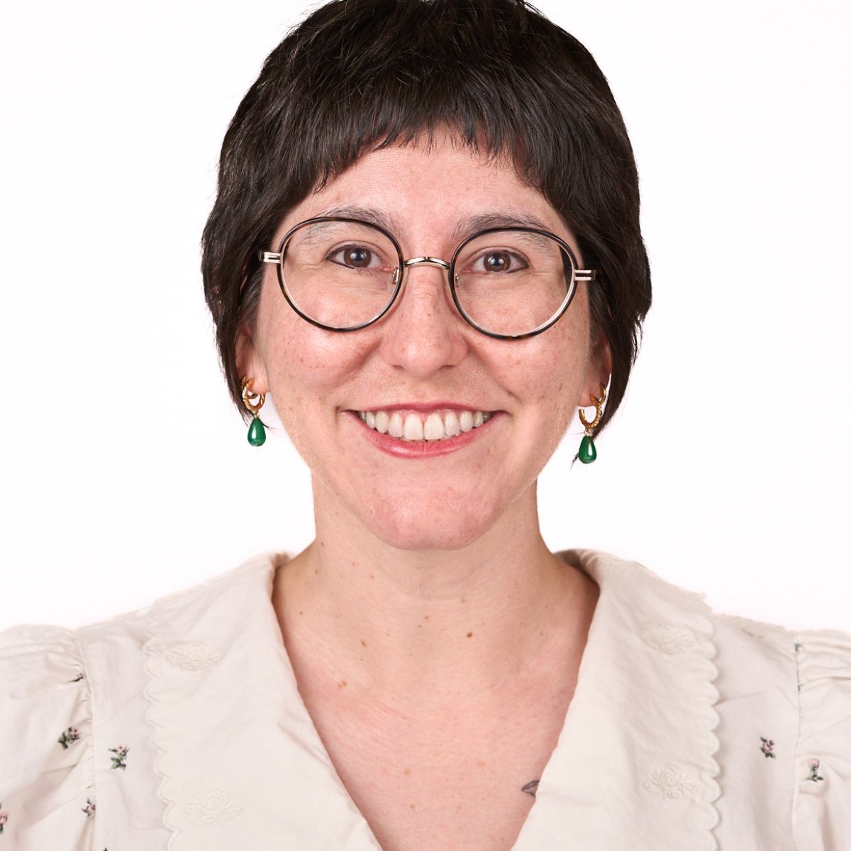"A smiling person with short dark hair, round glasses, and green drop earrings, wearing a light-colored blouse with floral embroidery, posed against a plain white background."