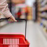 Female Hand Holds Empty Red Grocery Cart, Close-up. In Backgroun