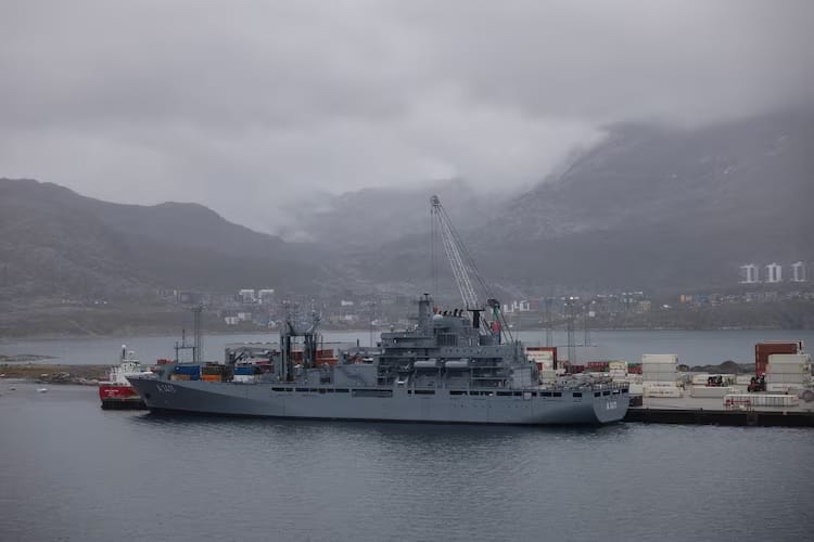 A large gray naval ship docked at a port in Greenland, with cranes and shipping containers nearby. Misty mountains and low clouds rise in the background, giving the scene a cold, overcast atmosphere.