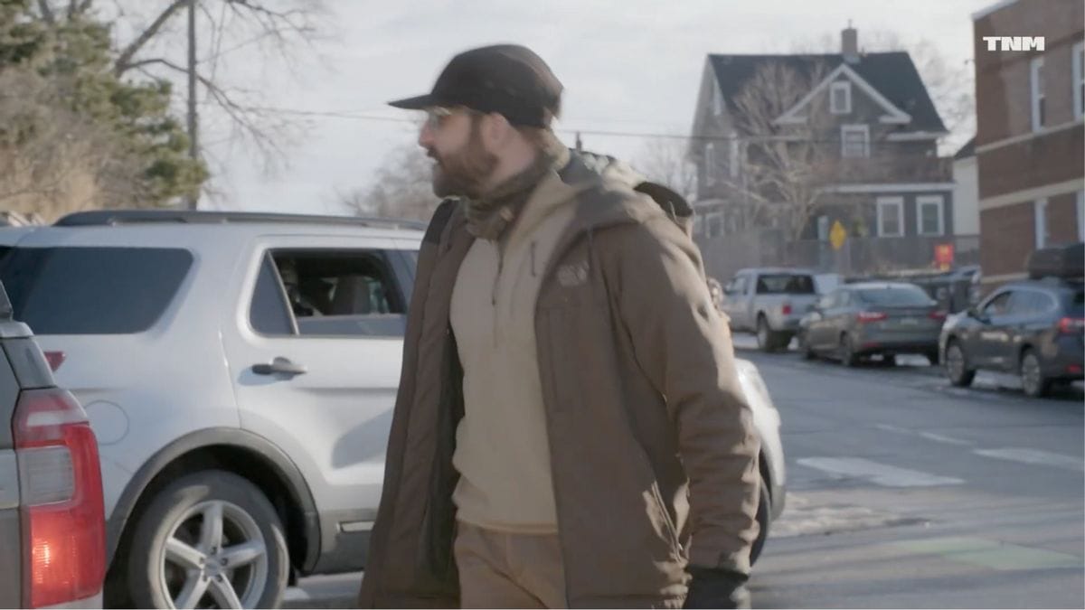 A slightly blurry image shows a white man wearing glasses, a hat and a hoodie speaking while looking to the left. He stands in a street, with cars parked near him and on the other side of the street. A white logo that says TNM can be seen in the top right corner.