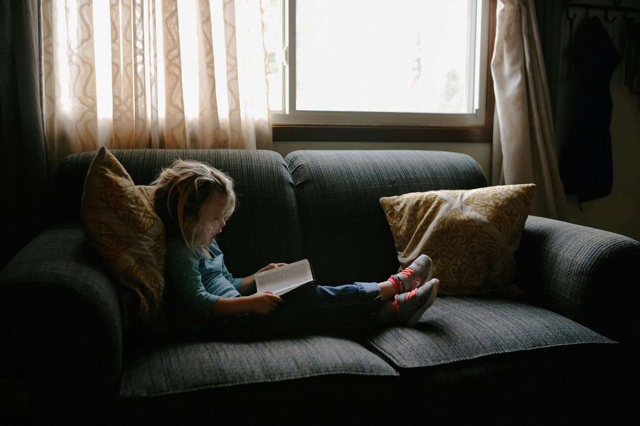 A child sitting on a grey couch by a window reading a book