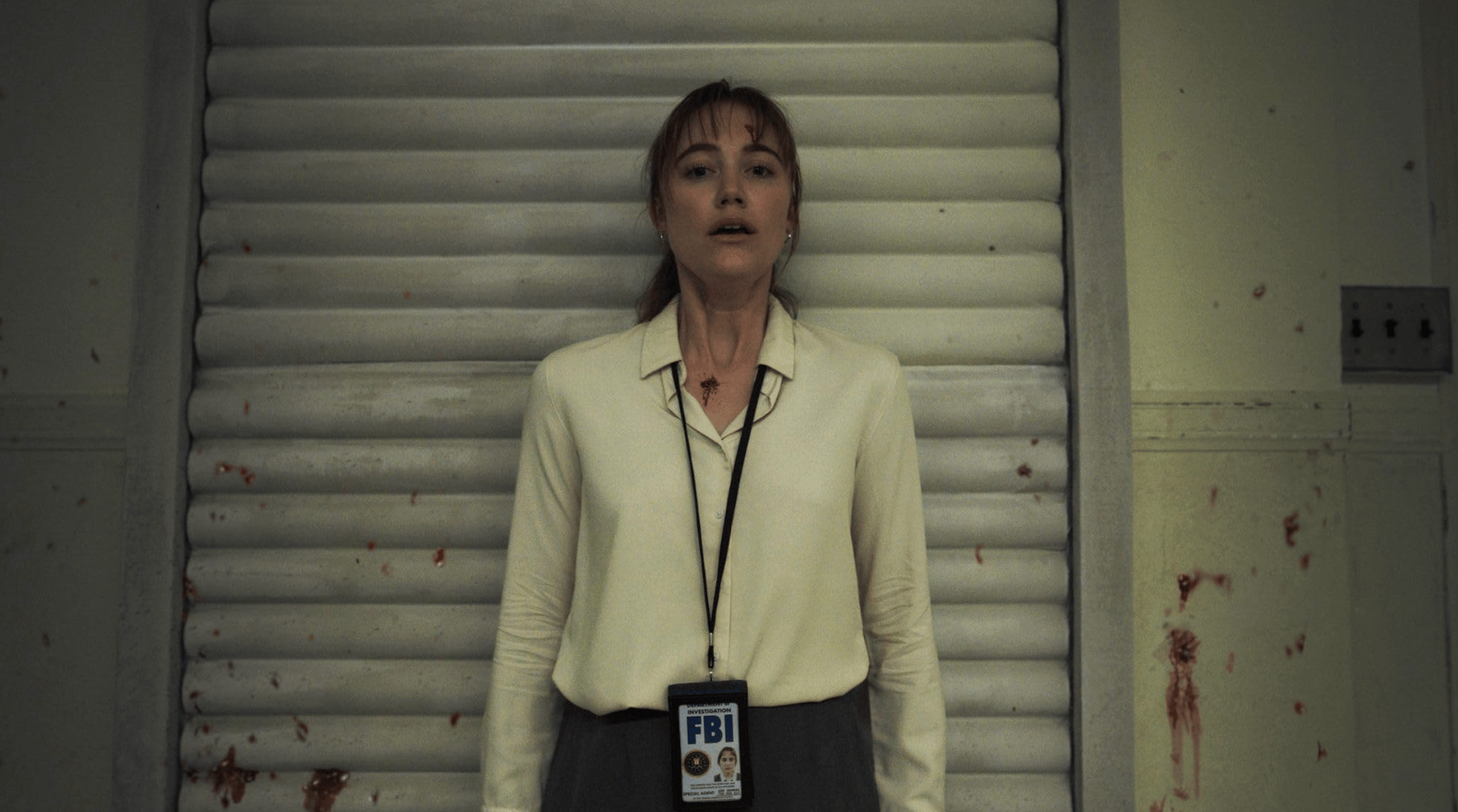 A white woman wearing an FBI badge standing against a blood-splattered wall with a look of shock on her face.
