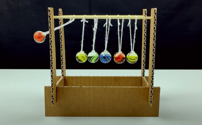 A photo of a homemade Newton's cradle made with cardboard, brightly-colored marbles and string. One ball is in motion and about to strike the row of stationary balls.