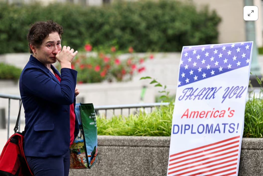 "A tearful person in a navy suit wipes their eyes while carrying bags, standing next to a sign that says 'THANK YOU America's DIPLOMATS!' decorated with an American flag motif, outside what appears to be a government building with flowers and greenery in the background."
