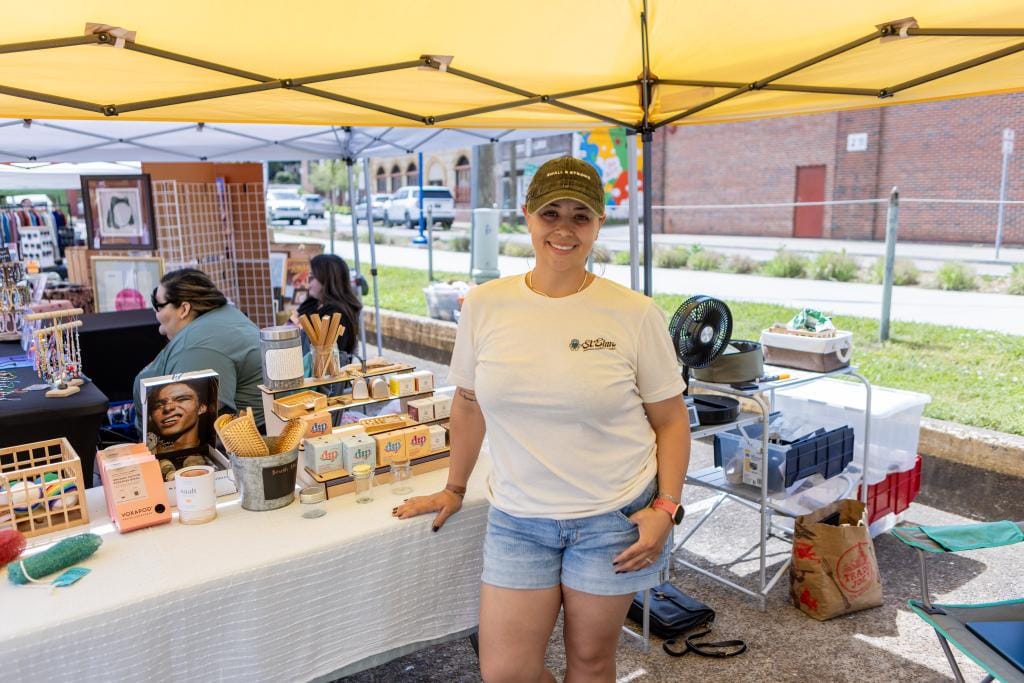 Tiffany from Full Circle Refillery stands smiling under a yellow canopy at a market, surrounded by sustainable goods and handmade products on display.
