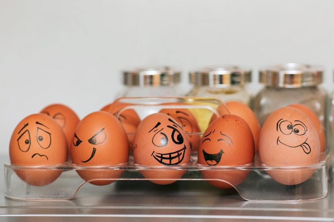 A photograph (wallpaper, picture) of eggs with different emotions against a background of blurred spices and a smooth gray wall. Whole painted eggs in the kitchen, expressing a spectrum of emotions from sadness to joy or madness. Eggs with smile and crazy face.