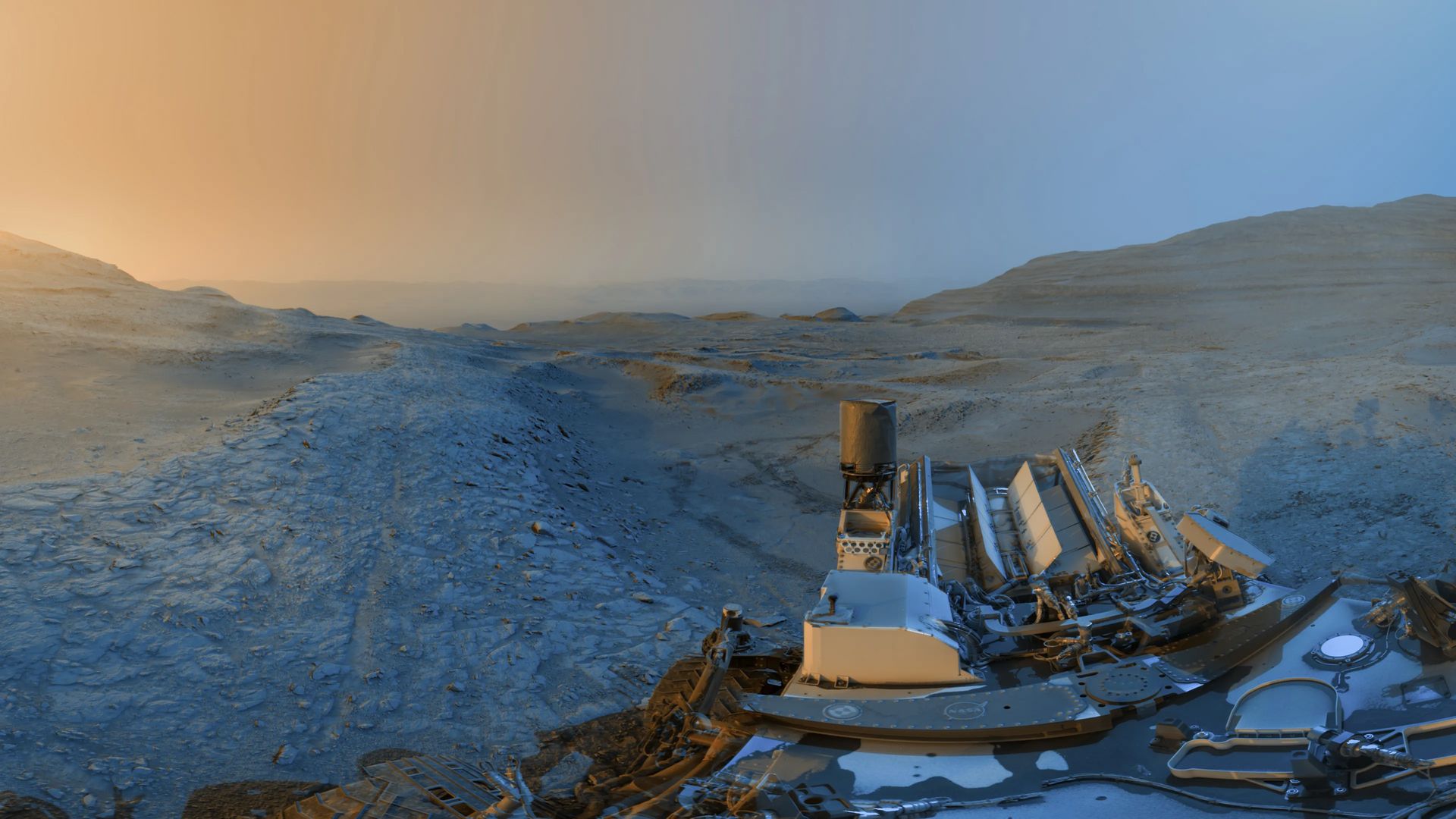 NASA’s Curiosity rover has released a new composite “holiday postcard” from its position on the slopes of Gale Crater, created by stitching together two panoramas taken on Nov. 18, 2025, during the rover’s 4,722nd and 4,723rd Martian days. The images were captured at 4:15 p.m. and 8:20 a.m. local Mars time, then merged to show the contrasting hues of a Martian afternoon and morning, blue tones from the early panorama and yellow from the later one, added as an artistic interpretation to highlight landscape details.
