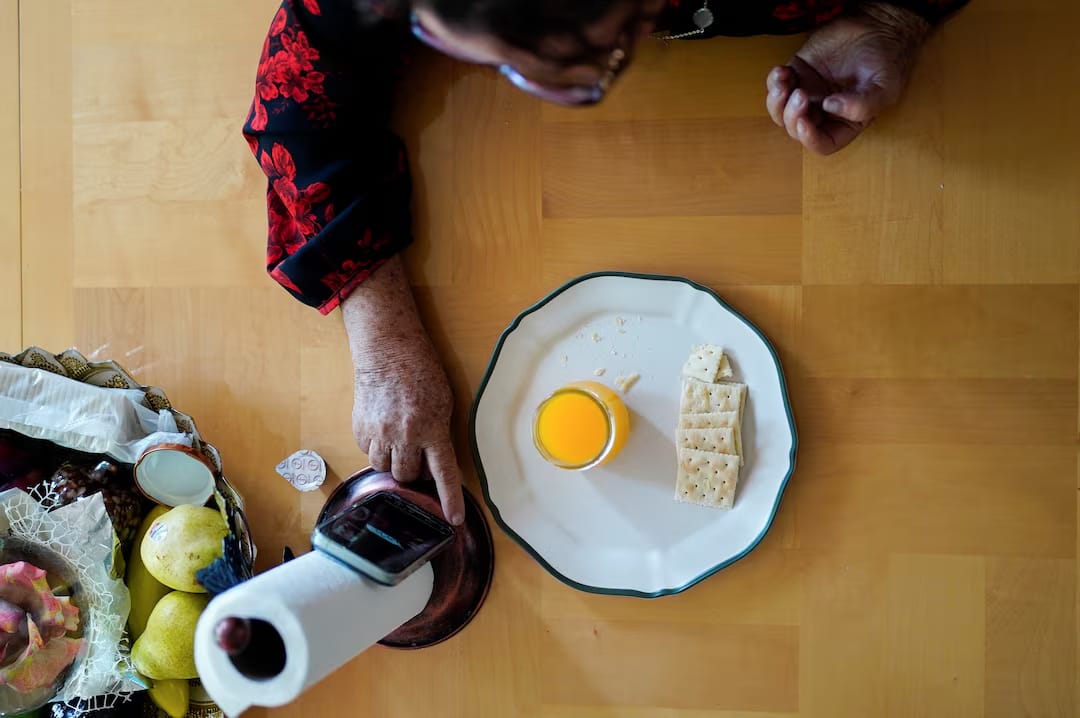 An overhead view of a woman sitting at a wooden table with a plate holding several crackers and a small glass of orange juice for home communion, watching a virtual church service on her phone propped up on a paper towel holder.