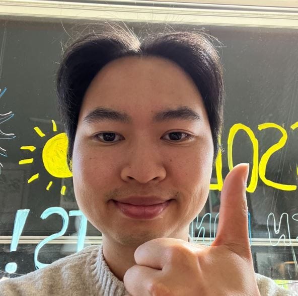 A person in a beige sweater gives a thumbs-up, standing in front of a window with a yellow sun and blue sky drawing, and "SOME" written in yellow.
