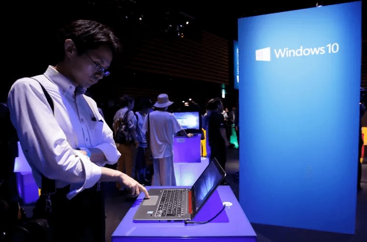 A man using a laptop at a Microsoft event with a large Windows 10 display stand in the background and other attendees around him. Select 76 more words to run Humanizer.