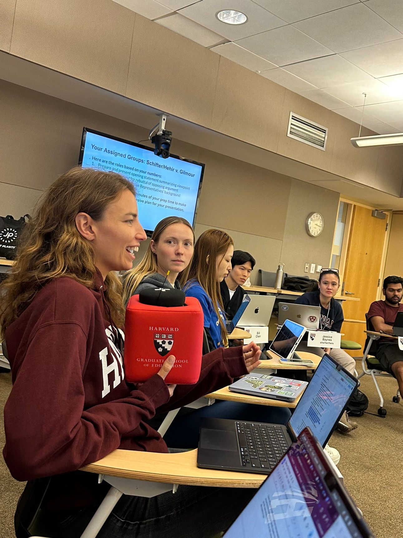 Image shows students in class at HGSE, with Tanyella in a red Harvard sweater speaking into a microphone, while her classmates listen. 