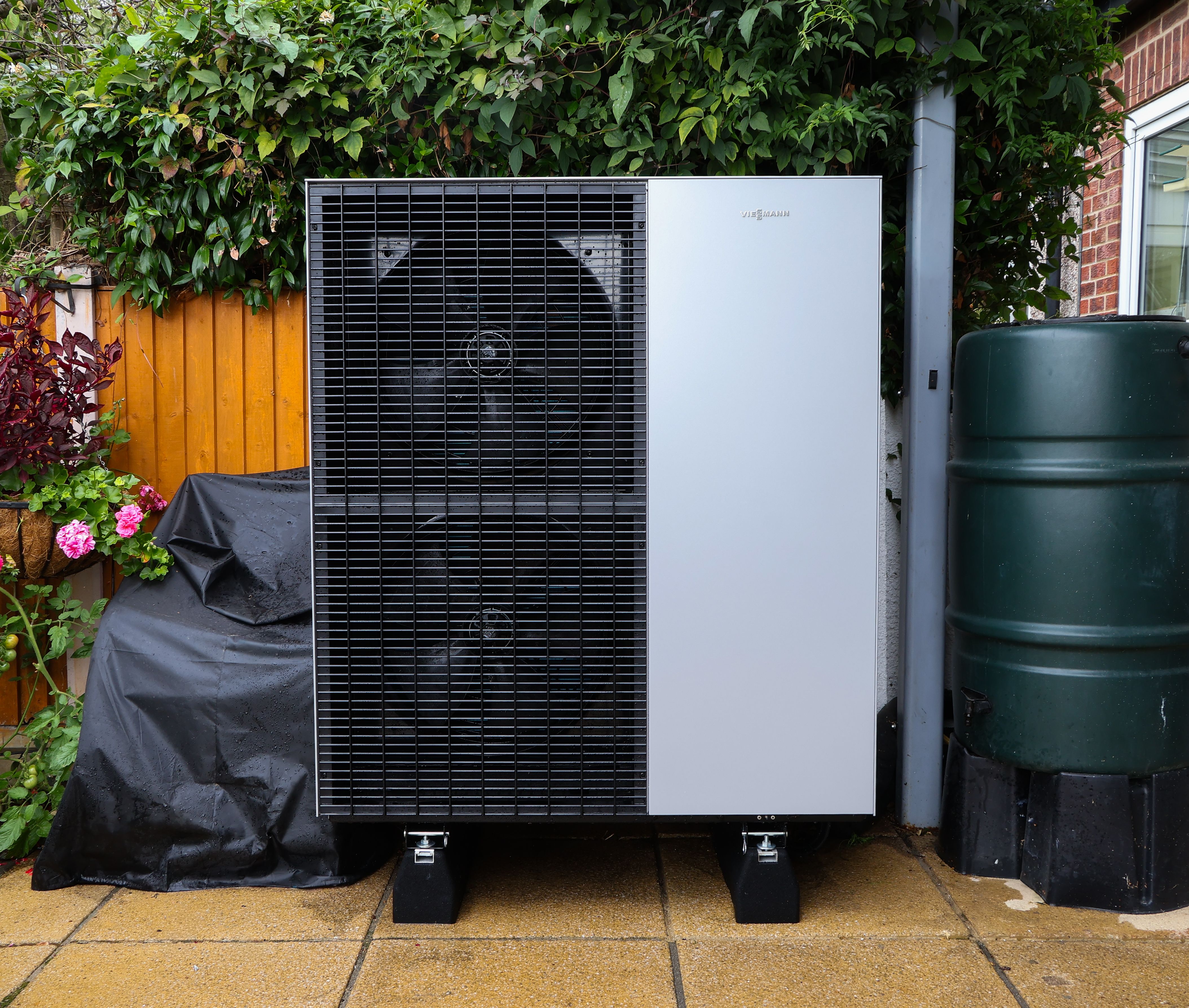 Close-up of a Viessmann Vitocal 150-A 13kW air source heat pump outdoor unit installed on anti-vibration mounts on a paved surface next to a garden water butt.
