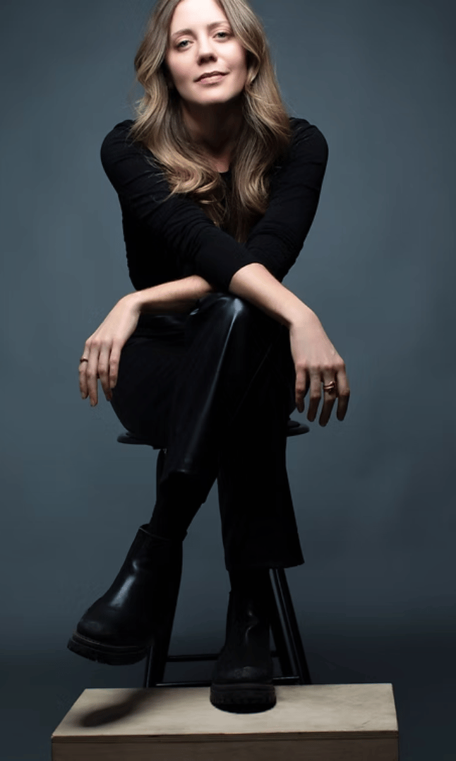 Bonnie Blue Edwards sits confidently on a black stool against a dark gray backdrop, dressed in black leather pants, a black long-sleeve top, and chunky black boots. Her arms are loosely crossed over one knee, with a calm and self-assured expression and soft waves in her long, blonde hair.