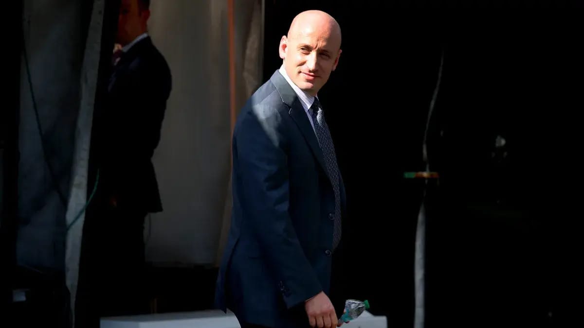 A white man wearing a blue suit stands looking slightly to the left. You can see another person standing slightly behind him.