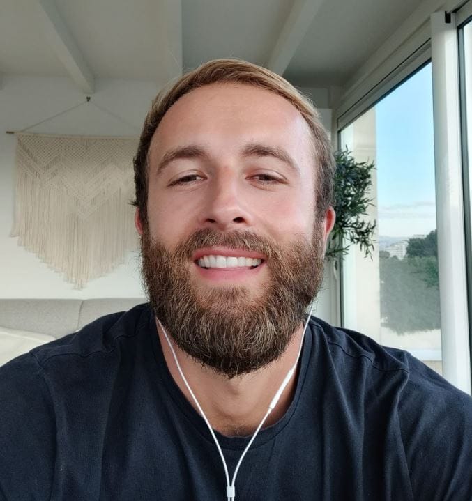 A man with a beard smiling at the camera, wearing a dark t-shirt and white wired earbuds.