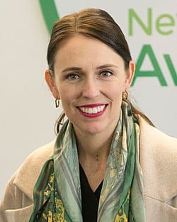 A woman with dark brown hair tied back, wearing gold hoop earrings, a patterned green scarf, and a light beige coat smiles at the camera against a blurred background with green text.