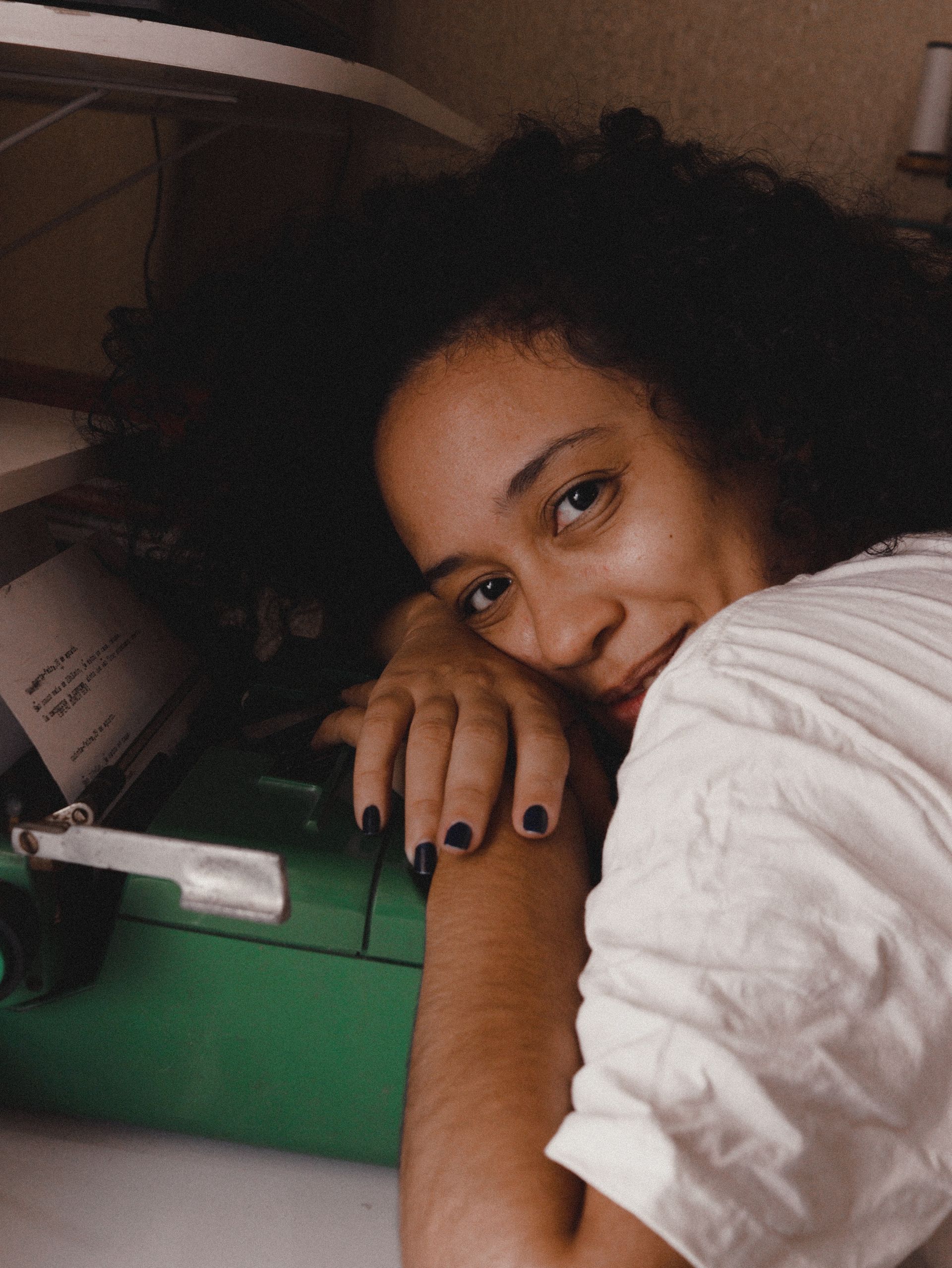 Fotografia da autora que mostra uma mulher negra de cabelos pretos cacheados. A autora se apoia numa maquina de escrever, com a cabeça reclinada sobre as mãoss, e sorri para a câmera.