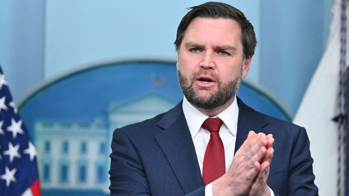 A white man wearing a blue suit stands with his hands pressed together while speaking. 