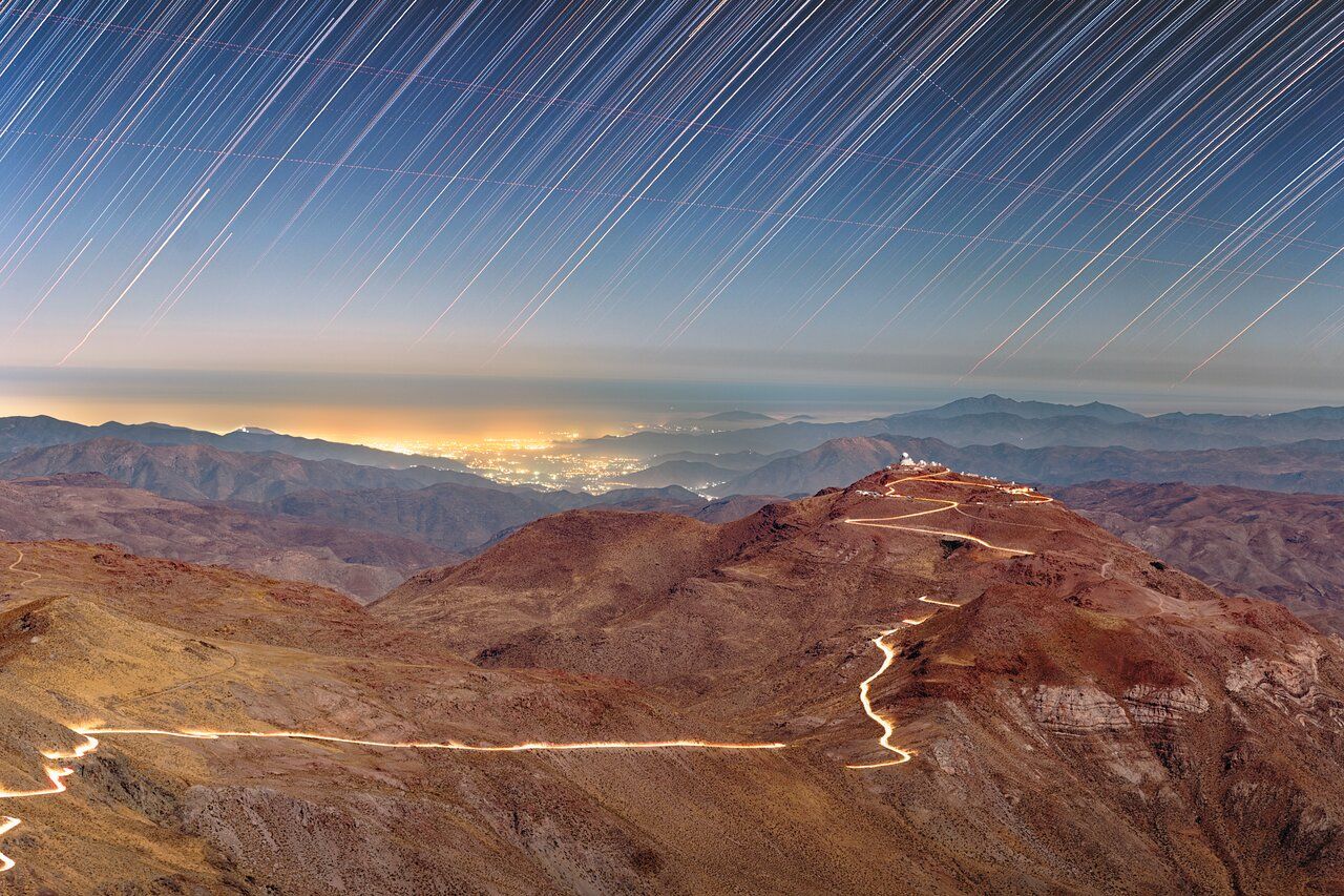 High in the Chilean Andes, two neighboring mountaintops host some of the world’s most advanced observatories. This long‑exposure image from Cerro Pachón, a site that also hosts the Vera C. Rubin Observatory, Gemini South, and the SOAR Telescope, captures the illuminated road linking Cerro Pachón to Cerro Tololo, its glow created by headlights of vehicles moving between the two sites. Cerro Tololo is home to roughly 40 instruments of the Cerro Tololo Inter‑American Observatory. Light from nearby towns, passing aircraft, and setting stars adds context, illustrating how these observatories operate within broader environmental and community landscapes of the Atacama Desert. 