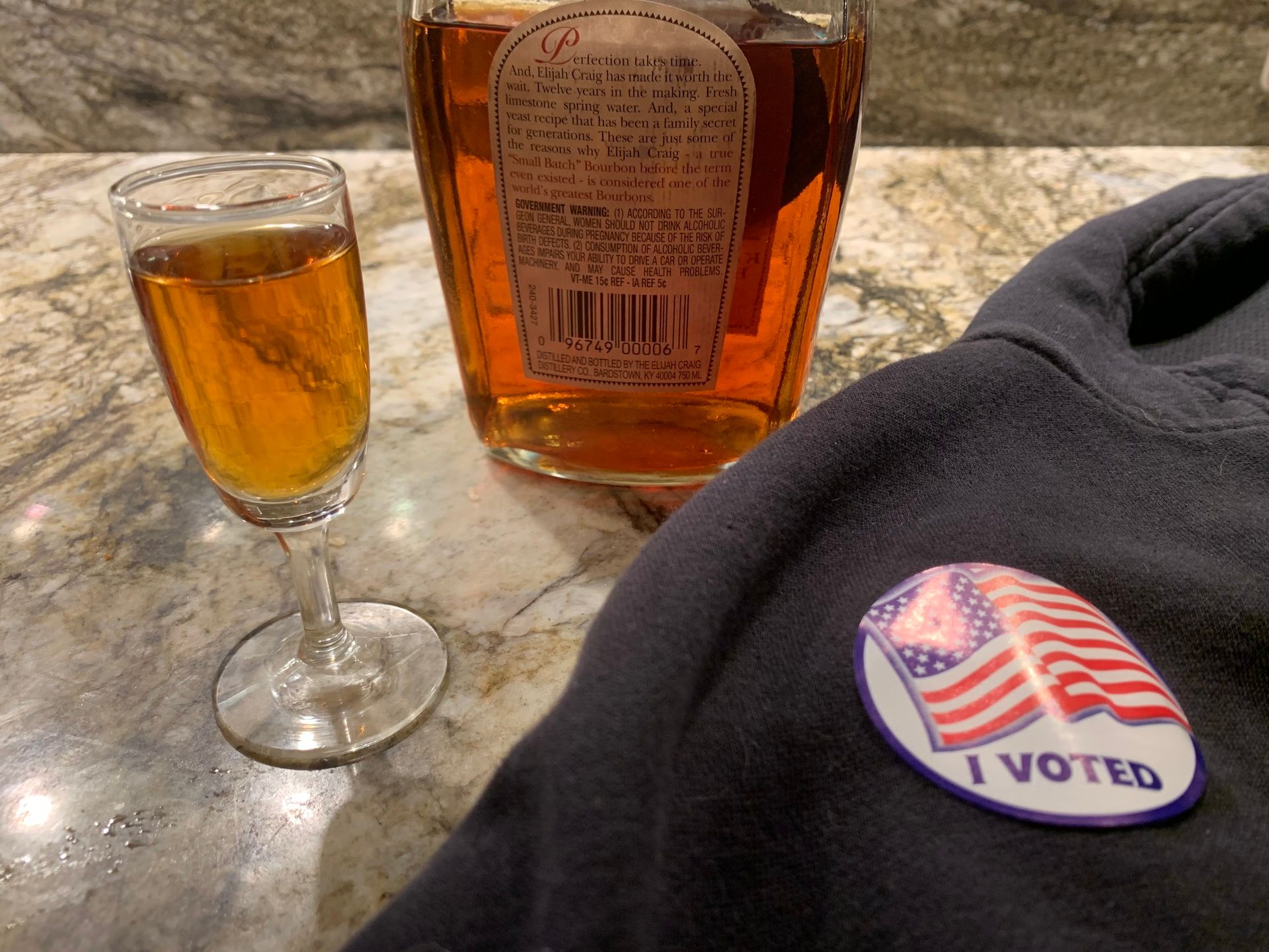 A photo of a bottle of bourbon, a small flute shot glass filled with liquor, and an "I VOTED" sticker on a hoodie.