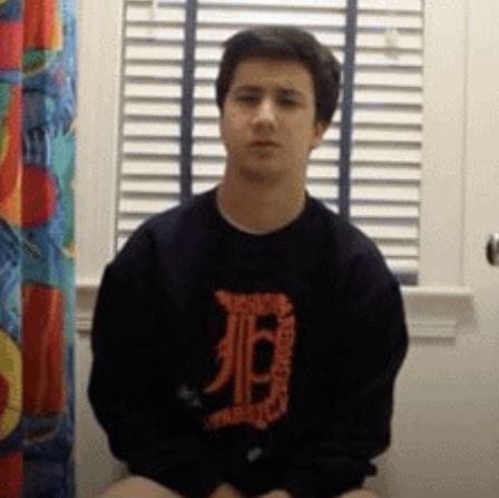 A young man in a black sweatshirt with a red logo sits in a room with colorful curtains, a white door, and closed blinds. The setting appears casual and indoor.