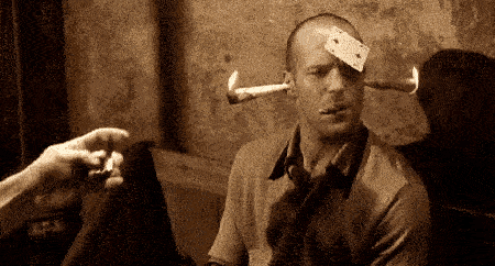Jason Statham in a pub with friends. He has a card stuck to his forehead, cigarette in mouth, and lit matches stuck in his ears.e