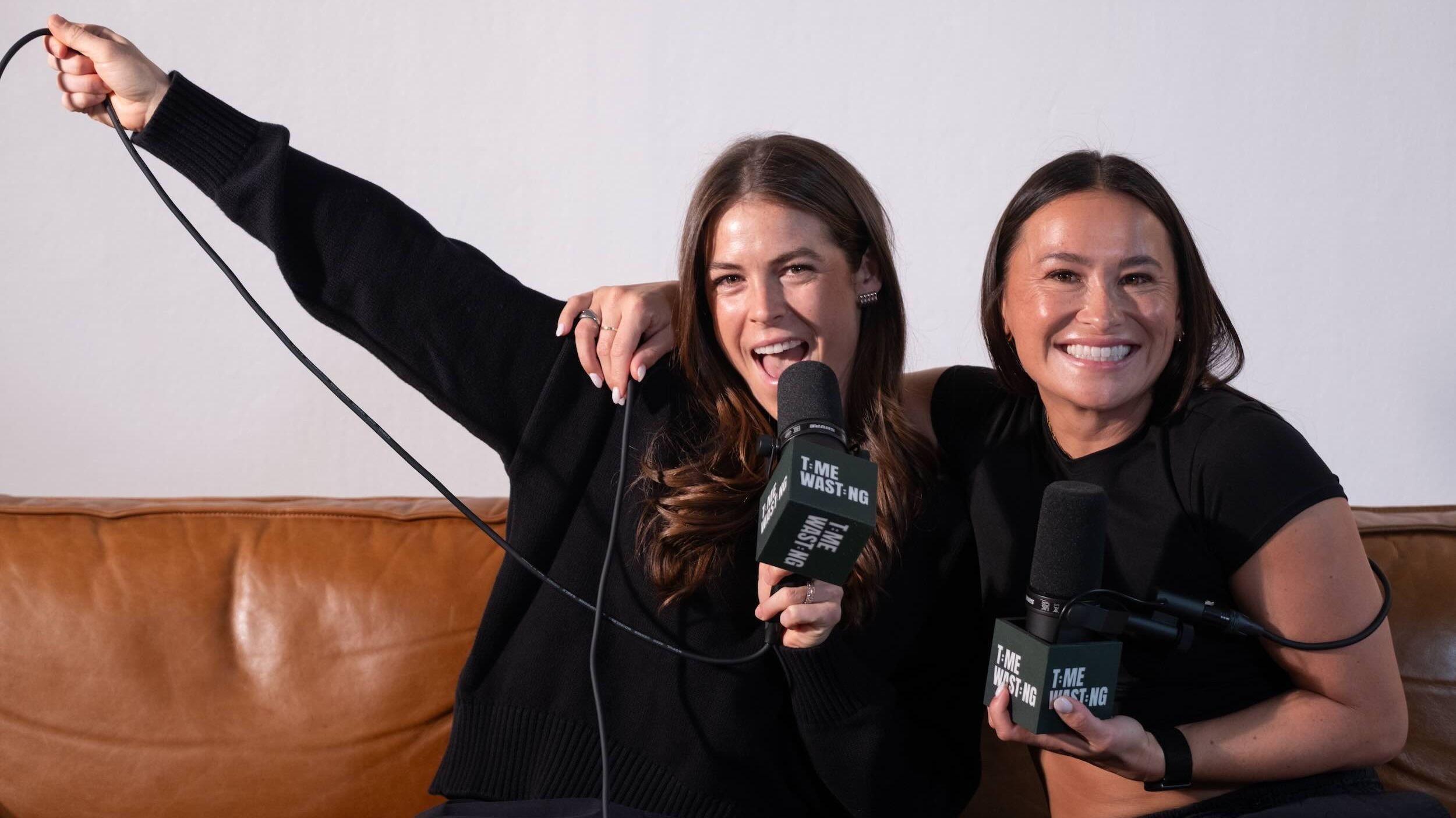Kelly O'Hara and Ali Riley pose on a couch with Time Wasting-branded mics.