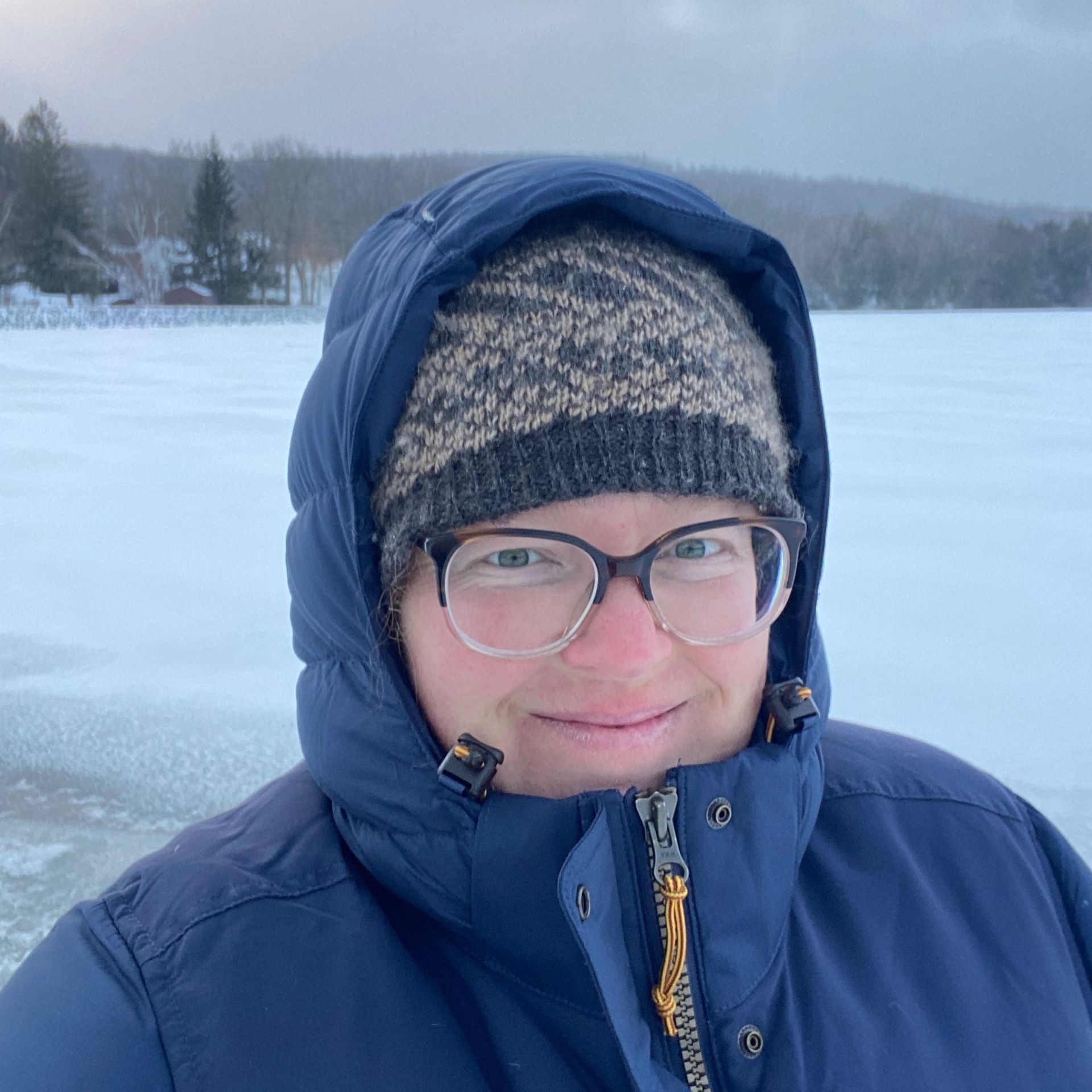 I’m standing in front of a frozen lake on a gray winter day in a puffy coat and wool hat.