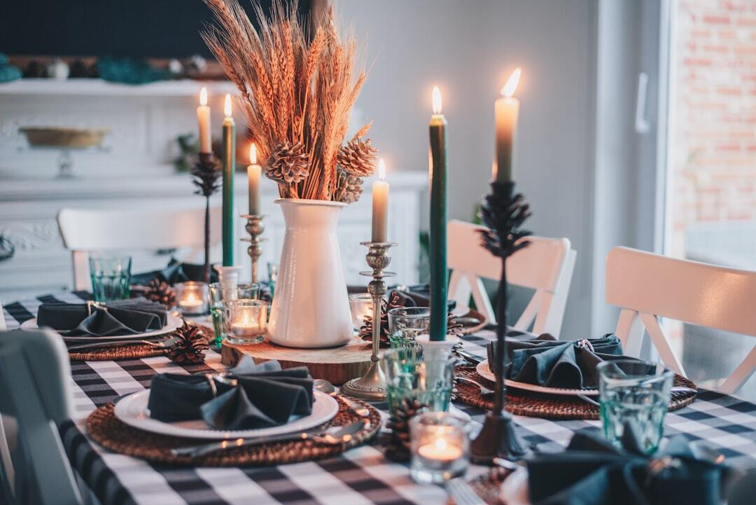 A dining table set with a black and white gingham tablecloth, plates with dark cloth napkins folded intricately on top, green lit candles, and a vase of wheat.