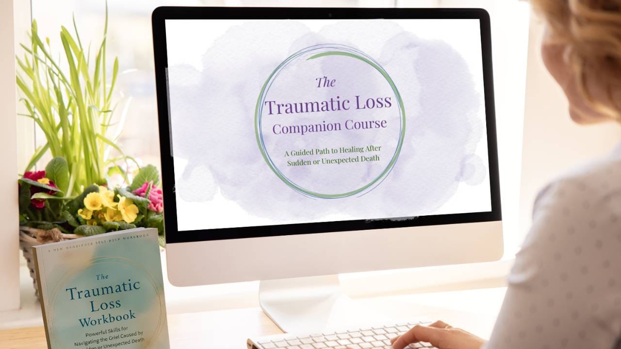 A woman sits at a desk looking at a computer screen displaying the title slide for The Traumatic Loss Companion Course: A Guided Path to Healing After Sudden or Unexpected Death. Beside the monitor, a copy of The Traumatic Loss Workbook rests on the desk. A window box with colorful flowers and tall green plants brightens the scene.