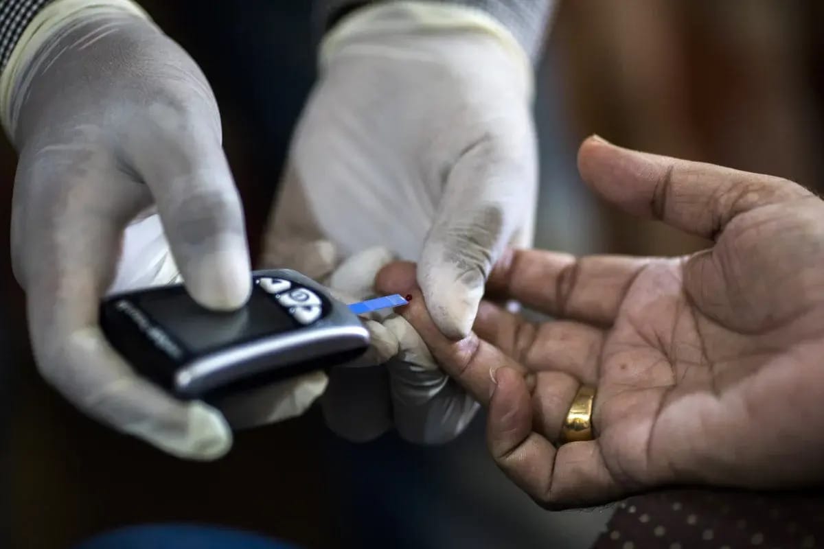 A healthcare worker wearing gloves uses a blood glucose meter to test a patient’s finger, where a small drop of blood is visible on the test strip.