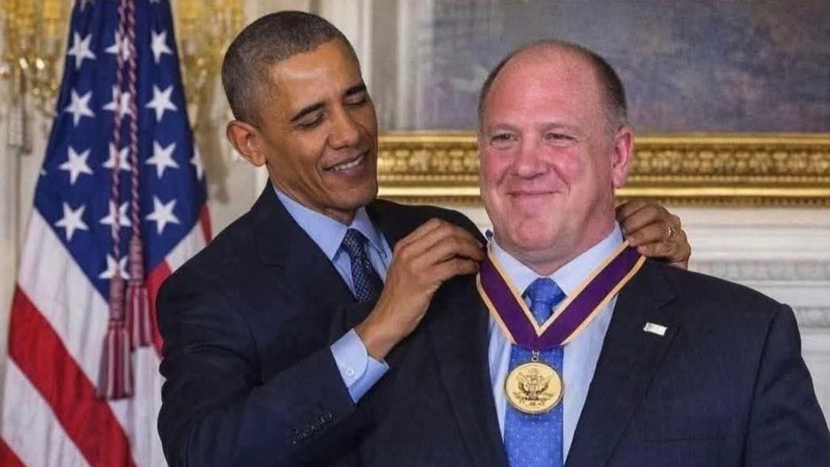 A Black man smiles as he places a medal around the neck of a white man who is also smiling.